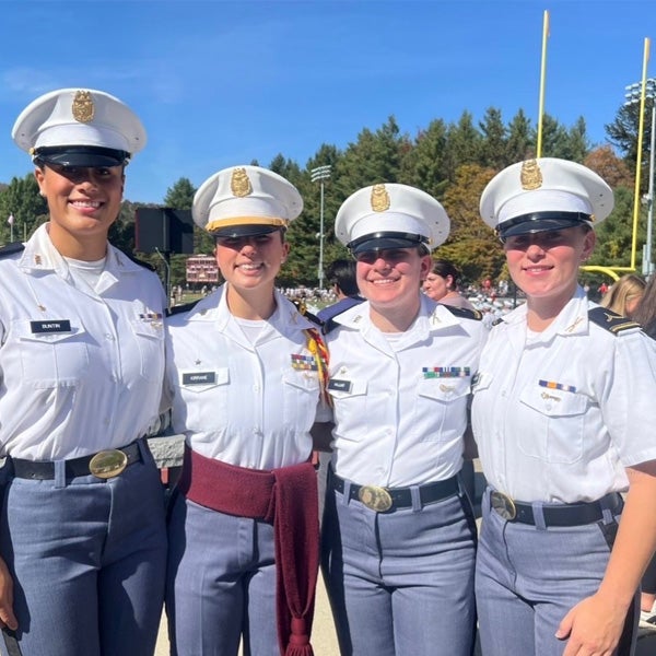 Four cadets in white dress uniforms stand together beside a football field on a sunny day.