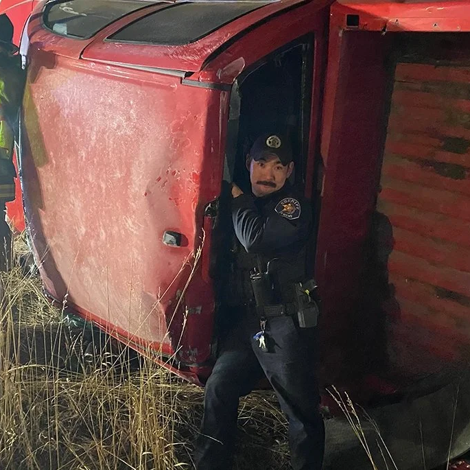 Police officer stands beside the open door of an overturned red vehicle at a nighttime crash scene.
