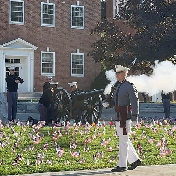 Ceremonial cannon firing by uniformed individuals in front of a brick building, surrounded by a field of small flags.