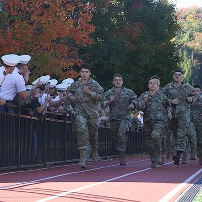 Service members in camouflage run on a track as uniformed spectators watch from behind a railing.