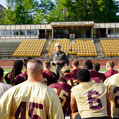 Football players gather around a coach speaking on the field in front of stadium seating.