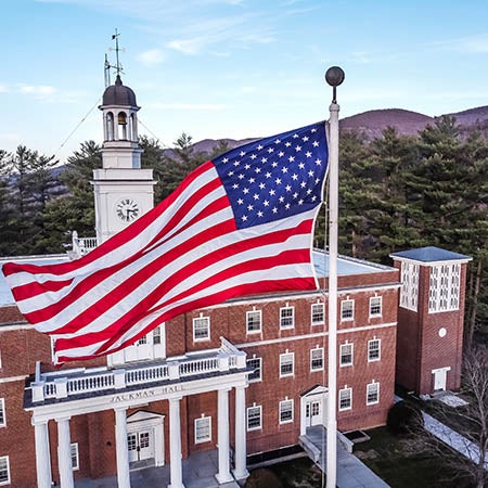 An American flag flies in front of a brick building with a clock tower and wooded mountains in the background.