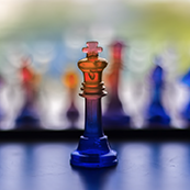 Orange and blue chess king stands on a board with blurred chess pieces in the background.