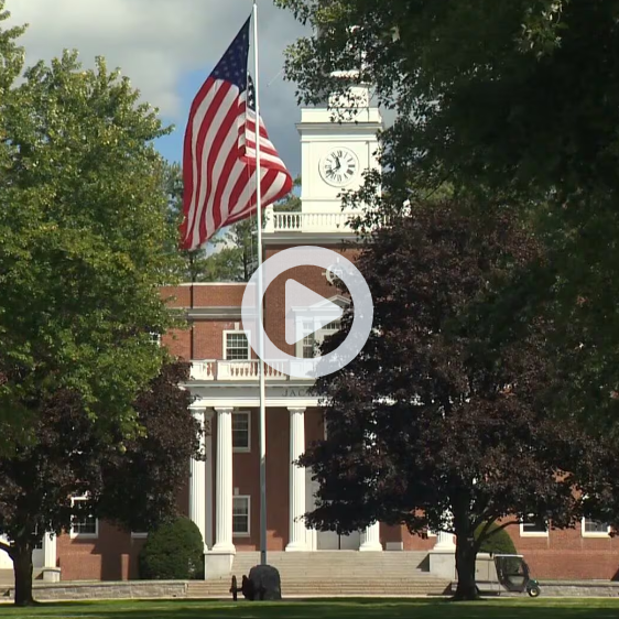 American flag flies in front of a brick building with white columns and a clock tower.