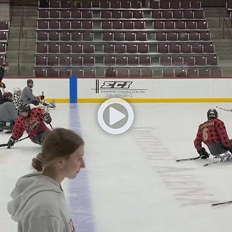 Sled hockey players practice with sticks and pucks on an indoor ice rink.
