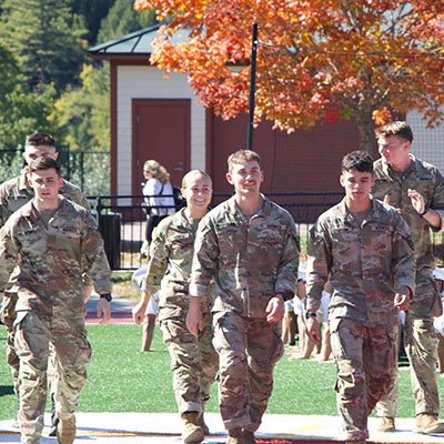Cadets in camouflage uniforms walk across an athletic field as other cadets exercise behind them.