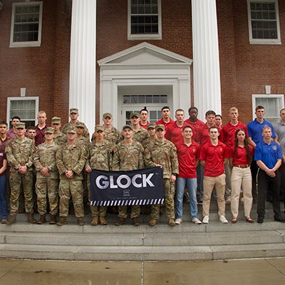 Cadets in camouflage uniforms and students in colored polo shirts pose with a GLOCK banner on steps outside a brick building.
