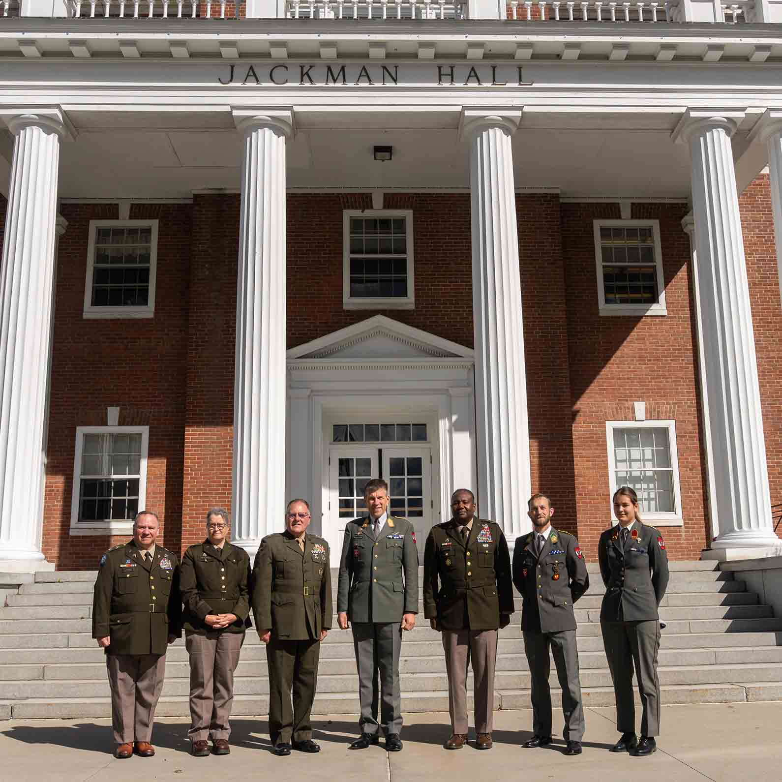 Seven service members in military dress uniforms stand on the steps in front of Jackman Hall at Norwich University.