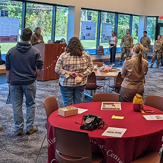 People stand around tables during a presentation as service members line the back of the room.