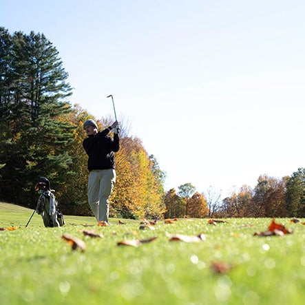 A golfer swings a club on a grassy course with autumn trees in the background.
