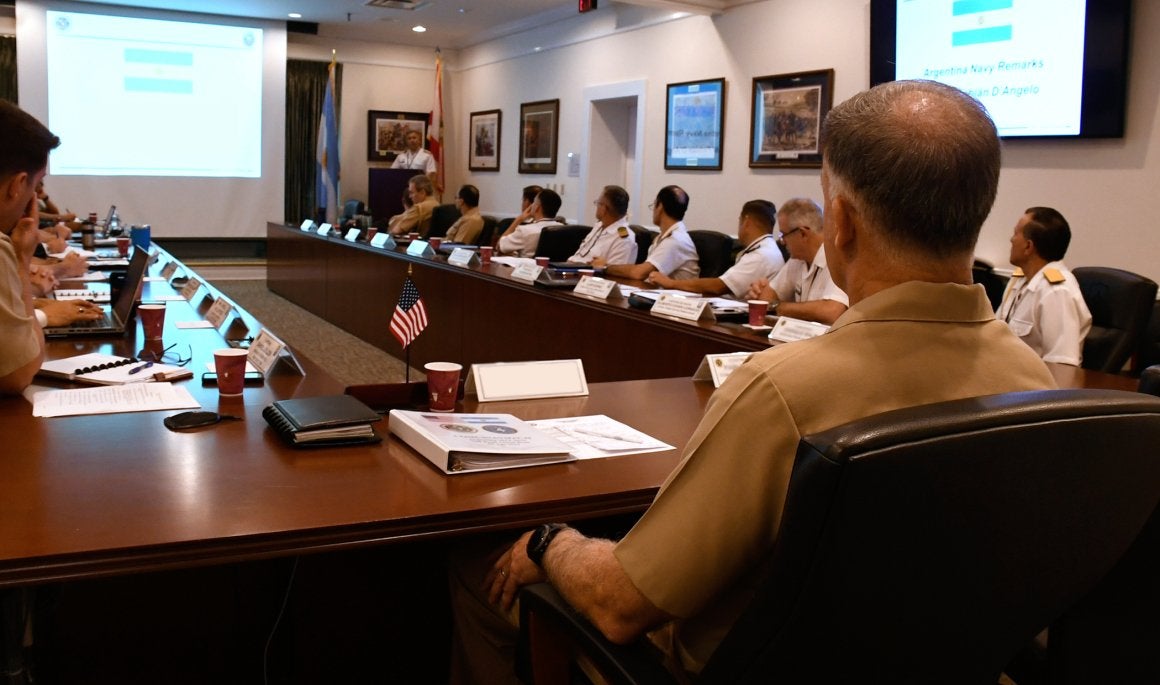 Military officers attend a conference room presentation with slides.