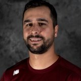 A man with short dark brown hair and a beard, wearing a maroon athletic shirt with a white logo, standing against a textured dark gray background.
