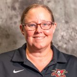 A smiling woman with short light brown hair, glasses, and a black polo shirt with a red and gold sports logo, standing against a textured gray background.
