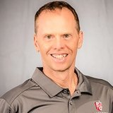 A smiling man with short brown hair wearing a gray polo shirt with a red and white logo, standing against a light gray background.