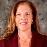 A smiling woman with long, wavy auburn hair, wearing a maroon blazer and gold jewelry, standing against a textured gray background.