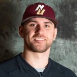 A man wearing a maroon baseball cap with gold and white initials and a dark zip-up jacket, smiling in front of a textured gray backdrop.