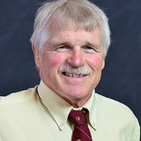 An older man with light gray hair and a mustache, wearing a pale yellow dress shirt and a maroon tie with a subtle pattern, smiling in front of a dark background.