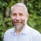 Man in a light blue shirt, smiling in front of foliage.