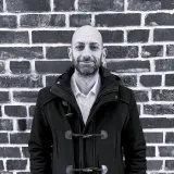 Man in a dark coat standing in front of a brick wall.