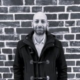Man in a dark coat standing in front of a brick wall.