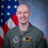 Smiling man in a green flight suit stands in front of a U.S. flag, with Air Force insignia and a name patch reading “Ryan Brown” on his uniform.