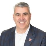 A man with short gray hair wears a plaid jacket, white shirt, and an N logo pin, standing against a white background.