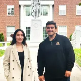 Two people stand outside near a statue in front of a red brick building with white columns.