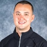 A person with short dark hair wears a dark zip-up jacket and smiles at the camera against a mottled gray background.