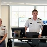 Two cadets in uniform are standing at the front of a classroom giving a presentation. One is speaking while the other smiles and listens. Both are wearing white shirts with rank insignia and ribbons. Behind them, a screen displays a slide with “ROTC Scholarship Requirements” listed. The environment appears professional and academic, with computers, notes, and coffee cups visible on the desks.