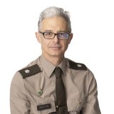 Person in a tan military-style uniform with shoulder insignia, name tag reading “MARTIN,” tie, and pins on the chest.