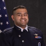 Service member in dark dress uniform stands in front of a U.S. flag and dark backdrop.