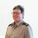 Person wearing glasses and a gray uniform shirt with shoulder insignia poses against a plain light background.