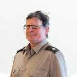 Person wearing glasses and a gray uniform shirt with shoulder insignia poses against a plain light background.