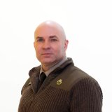 Person in a dark zip-up sweater and tie faces the camera against a white background.