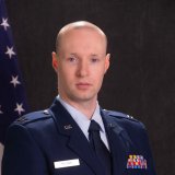 Uniformed service member stands in front of a U.S. flag and dark backdrop.