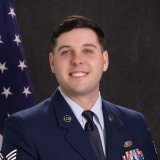 Service member in blue dress uniform stands in front of a U.S. flag and dark backdrop.