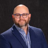 Gregory Mcgrath profile image with blue glasses and a beard wears a blue blazer and checkered shirt against a dark backdrop.