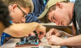 Two Norwich University students work with focused concentration, assembling electronic components on a circuit board at a classroom table.