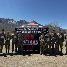The Norwich University cadets who participated in the event stand shoulder-to-shoulder in front of a branded tent in the desert with a mountain range in the background.