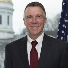 A formal headshot of Governor Phil Scott in a suit flanked by an American flag behind his left shoulder, and the capitol building in the distance behind his right.