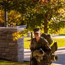 A cadet walks along a sidewalk in full uniform with a pack while smiling at the camera.
