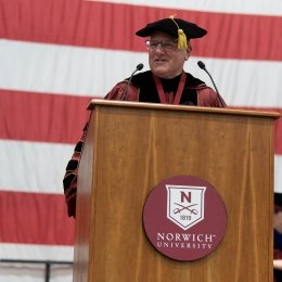A person in academic regalia delivering a speech at a podium with the Norwich University logo, in front of a large American flag.