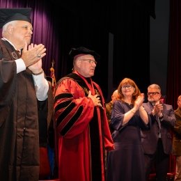John J. Broadmeadow, University president, stands in the center of the stage in full academic regalia with hand over heart. Chairman Alan DeForest is on his right, while his wife Karen is on his left.