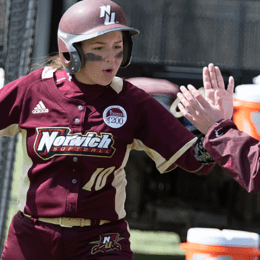 Norwich softball player in maroon uniform and batting helmet receiving high-five from coach near dugout, wearing number 10 jersey.