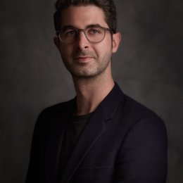 Portrait of an individual with glasses wearing a dark blazer, set against a textured grey background.