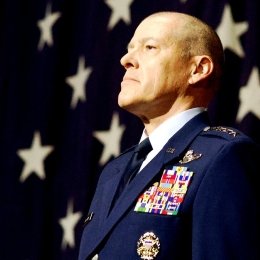 General Bussiere stands at attention in full dress Air Force uniform in front of a large American flag.