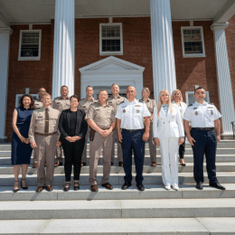 Group of uniformed military personnel and civilians posing together on steps in front of a building with large columns.