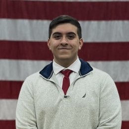 A portrait headshot of Hector Aponte in a sweater, shirt and tie in front of American flag.
