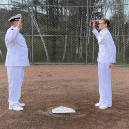 Caitlin Dailey (right) stands next to home plate while her father (left, directly across home plate) swears her in. They are both in full white Navy outfits.