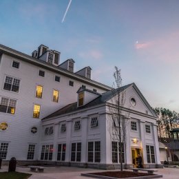 Large white building with square windows and lit rooms at dusk, with clear sky and trees in the background.